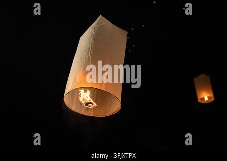 Warm leuchtende Papierhimmelslaterne steigt nachts auf mit sichtbarer Flamme im glühenden dünnen Papier und verschwommenen Laternen im Abendhimmel im Freien Stockfoto