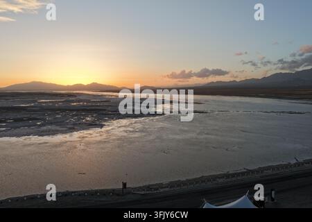 Eine tiefe Sonne versinkt hinter sanften Bergrücken und wirft warmes Licht und lange Silhouetten über eine ruhige Salz Ebene. Das friedliche Hochlandbecken leuchtet im Abendlicht Stockfoto