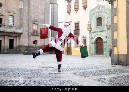 Lustiger Weihnachtsmann, der auf einem Bein balanciert und während der Weihnachtszeit farbenfrohe Weihnachtseinkaufstüten auf einem historischen Stadtplatz hält. Stockfoto