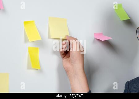 Junger männlicher Büangestellter, der Projektideen auf Klebezetteln brainstormt und auf eine Bürowand schreibt. Stockfoto