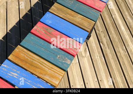 Holzlatten bilden eine helle, abgenutzte Bank, die eine lebendige Auswahl an gemalten Farben und gealterten Texturen zeigt, die auf einem sauberen Holzdeck ruht unter Stockfoto