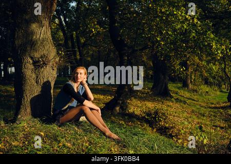 Nachdenkliche Frau sitzt auf dem Gras neben einem Baum im Park. Das Licht der untergehenden Sonne fällt auf ihr ruhiges Gesicht Stockfoto
