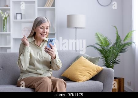 Zufriedene Seniorin auf einem Sofa feiert gute Nachrichten auf ihrem Smartphone, lächelnd vor Begeisterung in einem hellen modernen Wohnzimmer, entspannt und den Moment genießend Stockfoto