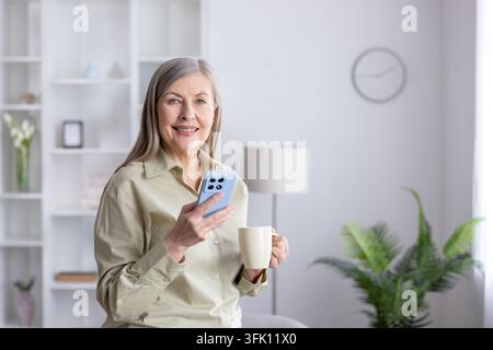Reife Frau, die aktiv mit ihrem Smartphone interagiert und eine Kaffeetasse hält, die digitale Verbindung und modernen Lebensstil demonstriert, während sie in einer hellen häuslichen Umgebung lächelt Stockfoto