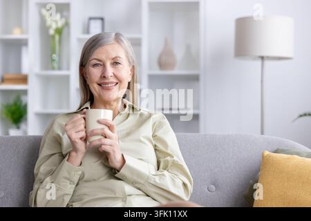 Seniorin mit grauen Haaren sitzt bequem auf einem grauen Sofa, lächelt fröhlich und hält eine warme Kaffeetasse in ihrem Wohnzimmer, genießt Komfort und Entspannung Stockfoto