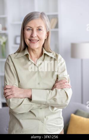 Reife grauhaarige Frau steht in einem hellen Heimbüro, lächelt mit verschränkten Armen und strahlt selbstbewusste, erfahrene, professionelle Wärme und zugängliche Eleganz aus Stockfoto
