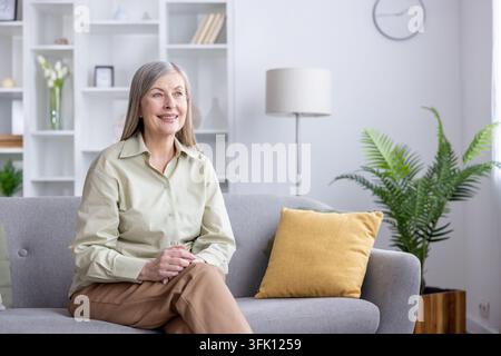 Seniorin sitzt bequem auf einer grauen Couch in einem hellen Wohnzimmer, lächelt friedlich und genießt einen ruhigen Moment der Entspannung und Zufriedenheit zu Hause Stockfoto
