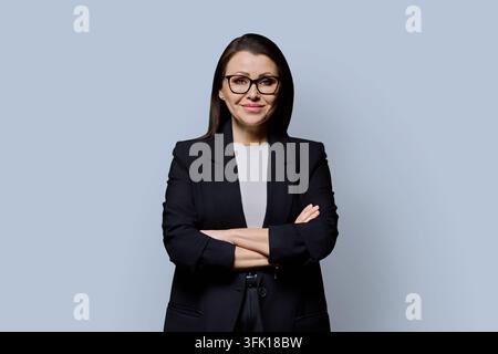 Lächelnde reife Geschäftsfrau mit verschränkten Armen trägt Brille vor grauem Hintergrund Stockfoto