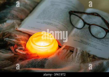 Brennende Kerze, Buch und Brille mit einer karierten Decke. Gemütliches Zuhause, Herbstkonzept, Nahaufnahme. Stockfoto