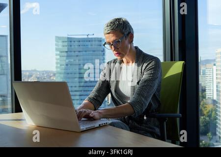 Geschäftsfrau, die an einem Laptop arbeitet, in einem modernen Büro mit Stadtblick Stockfoto