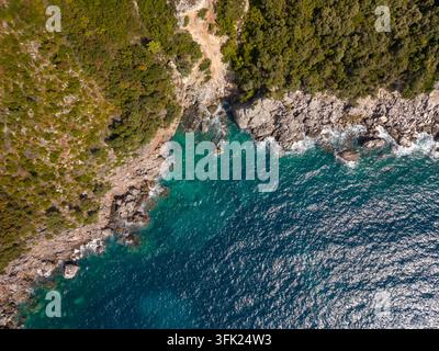 Felsenküste grenzt an eine türkise Bucht in Aquarium Bay, Albanien. Luftaufnahme zeigt klares Wasser vor steiler Küste und dichtem grünem Buschland, mit felsigem Innenland Stockfoto