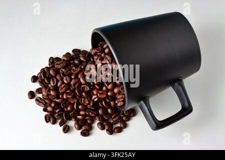 Mainz, Rheinland-Pfalz, Deutschland: Geröstete Kaffeebohnen, die aus einer schwarzen Keramiktasse auf sauberem weißem Hintergrund quellen. Stockfoto