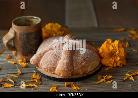 Stillleben mit traditionellem Pan de Muerto-Brot, einem kunsthandwerklichen Tonbecher und Cempasuchil-Goldblumen auf einem Holztisch Stockfoto