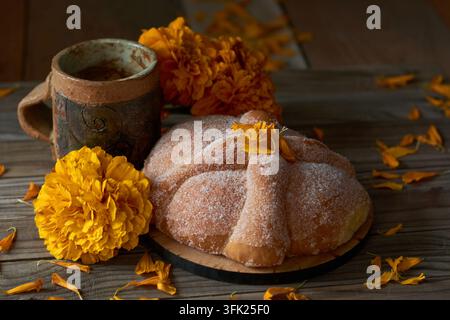 Stillleben-Variation des traditionellen Pan de Muerto-Brots mit Tonbecher und Cempasuchil-Marienkrautblüten auf rustikalem Holz Stockfoto