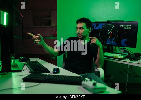 Attraktiver bärtiger gemischtrassiger männlicher Gamer sitzt am Schreibtisch, zeigt auf Bildschirme, die ein strategisches Simulationsspiel anzeigen, und verwendet Computerausrüstung im Raum Stockfoto