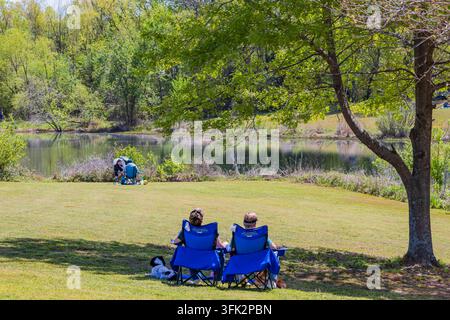 Arkansas, Vereinigte Staaten, 08. APR 2024 – Zwei Personen sitzen in blauen Klappstühlen auf einem grasbewachsenen Hügel mit Blick auf einen ruhigen See, der von üppigen grünen Bäumen umgeben ist. Stockfoto