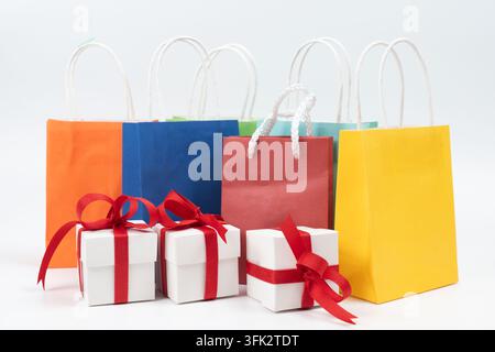 Bunte Einkaufstüten mit kleinen weißen Geschenkschachteln, die mit roten Bändern verziert sind. Hervorhebung von Feiertags-einkäufen, Einzelhandelsverkäufen, festlichen Rabatten, Geburtstagsfreuden und Verbraucherförderungskonzepten Stockfoto