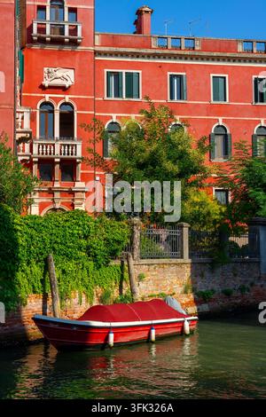 Rotes venezianisches Palais und ein festgemachtes Boot entlang eines ruhigen Kanals in Venedig . Stockfoto
