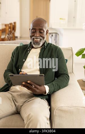 Senior Mann sitzt auf dem Sofa im Wohnzimmer und hält ein Tablet mit Hülle, trägt einen cremefarbenen Pullover Stockfoto