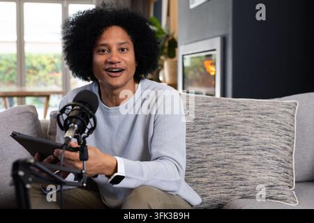 Afroamerikanischer Mann sitzt auf grauer Couch im Heimstudio und spricht in ein Mikrofon, während er ein Tablet hält Stockfoto