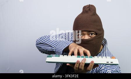 Ein erwachsener asiatischer Mann, der seine persönliche Identität mit einer braunen Sturmhaube verbirgt, beugt sich vor, während er aggressiv auf einer mechanischen Tastatur tippt. Der maskierte Individu Stockfoto