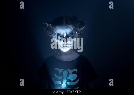 Kind mit Skelett-Gesichtsbemalung, von unten beleuchtet, das ein gruseliges Halloween-Porträt vor dunklem Hintergrund erzeugt. Stockfoto