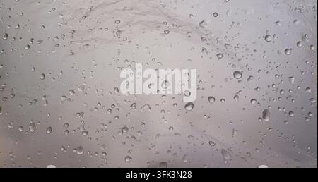 Regentropfen auf grauem nebligem Glas-Hintergrund mit weichem verschwommenem Licht hinter dem nassen Fenster. Stockfoto
