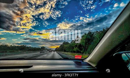 Die Aussicht aus einem Auto, das auf einer mehrspurigen Autobahn während eines dramatischen Sonnenuntergangs mit strukturierten, farbenfrohen Wolken fährt. Stockfoto