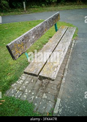 Eine alte, verwitterte Holzbank mit grünen Metallstützen, bedeckt mit Moos und Flechten, steht neben einem gepflasterten Weg und grünem Gras in einer Außenanlage Stockfoto