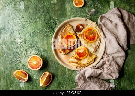 Hausgemachte Crêpes-Pfannkuchen serviert auf einem Keramikteller mit blutigen Orangen und Rosmarinsirup mit geschnittenen sizilianischen roten Orangen über grünem Texturrücken. Oberansicht, Platz Stockfoto