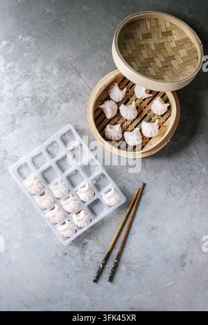 Gefrorene asiatische Knödel zum sofortigen Kochen, mit Garnelen gefüllt, in einer Plastikbox mit Essstäbchen und Bambusdampfgarer auf grauem Texturrücken. Oberansicht, Platz. Halbfertiges Produkt Stockfoto