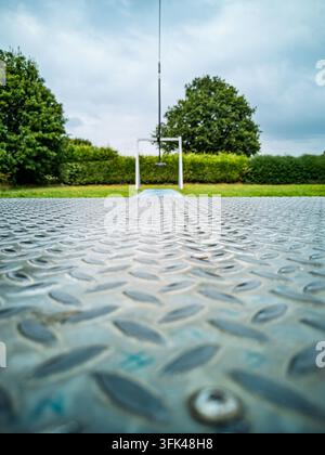 Eine niedrige Blickwinkelaufnahme über eine strukturierte Metall-Diamantplattenoberfläche hinweg zu einem grünen Park mit Hecken, Bäumen und einer weißen Metallkonstruktion unter einem bewölkten Himmel Stockfoto