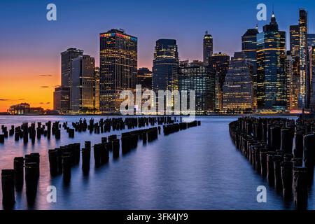 Ein atemberaubender Blick auf die Skyline von New York City vor einem lebendigen Sonnenuntergang. Reflexionen tanzen auf dem Wasser, während die Lichter der Stadt zu leuchten beginnen. Stockfoto