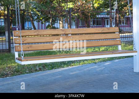 Leere hölzerne Schaukelbank, die durch Metallketten über einem gepflasterten Gehweg neben Bäumen aufgehängt ist. Stockfoto