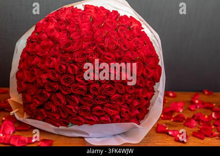 Großes Bouquet aus roten Rosen mit verstreuten Blütenblättern. Großes Bouquet dicht angeordneter roter Rosen, in weißes Papier gewickelt, ruht auf einem Holztisch Stockfoto