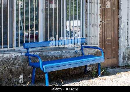 Eine verwitterte blaue Holzbank steht auf einem Bürgersteig vor einer verfallenen Gebäudefassade mit vergitterten Fenstern und einer rustikalen Holztür. Stockfoto