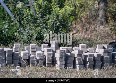 Mehrere Stapel grauer Betonpflastersteine stehen auf einem grasbewachsenen Straßenrand, der für ein Bauprojekt für einen Außenweg vorbereitet ist. Stockfoto