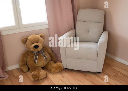 Teddybär sitzt in einem Raum mit einem Fenster in der Nähe des Stuhls Stockfoto