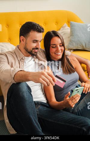 Ein lächelndes Paar sitzt eng beieinander auf einer gelben Couch, blättert auf einem Smartphone und bereitet eine Kartenzahlung für eine Online-Bestellung vor, was digitales Einkaufen und Stockfoto
