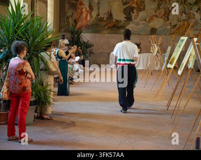 Cremona, Italien – 20. Juni 2026 Eine Gruppe von Menschen beobachtet eine Kunstausstellung in einem geräumigen Raum mit einem großen Wandgemälde und Topfpflanzen, während ein Ma Stockfoto
