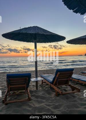 Leere hölzerne Sonnenliegen unter einem Strohdach, die einem atemberaubenden orangefarbenen Sonnenuntergang über dem Meer an der Strand von Phalasarna auf Kreta zugewandt sind. Stockfoto