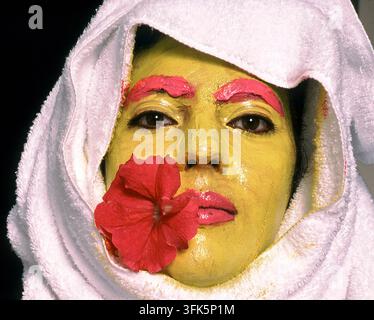 Eine Frau, die in ein weißes Handtuch gewickelt ist, trägt eine hellgelbe Gesichtsmaske mit rosa Make-up-Akzenten und einem roten Hibiskus auf ihrer Wange, was Schönheit und Entspannung ausstrahlt Stockfoto