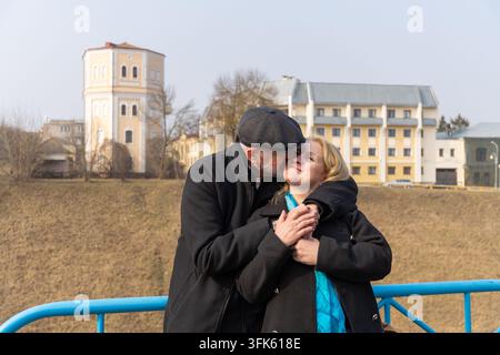 Romantisches reifes Paar umarmt sich, Mann küsst die Wange der Frau. Sie tragen schwarze Wintermäntel und einen blauen Schal in einer urbanen Außenszene mit historischen Gebäuden Stockfoto