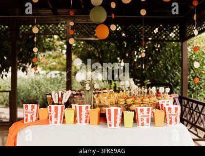 Tisch für Outdoor-Partys mit gestreiften Popcorn-Boxen, verschiedenen Snacks und hängenden Dekorationen, arrangiert für eine Feier oder ein Catering-Event. Stockfoto