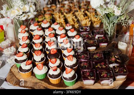 Erdbeer-bekrönte Cupcakes mit Sahneglasur und Schokoladen-Brownies, die auf Holzservierplatten für ein Dessertbuffet bei einer Veranstaltung angeordnet sind. Stockfoto