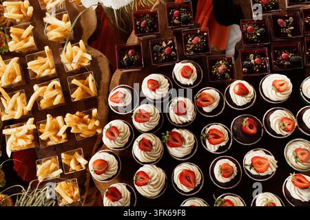 Oberblick über ein Partybuffet mit verschiedenen Cupcakes, Schokoladendesserts, Erdbeeren, Pommes frites und anderen Fingerfood, das für ein Eventcatering arrangiert ist Stockfoto