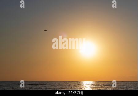 Ein kommerzielles Passagierflugzeug als Silhouette vor einer hellen goldenen Sonne, das bei Sonnenuntergang über den ruhigen Ozean fliegt. Stockfoto