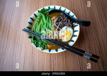 Hochwinkel-Ansicht von köstlicher asiatischer Ramen-Nudelsuppe mit gekochtem Ei, grünem Gemüse, schwarzem Morchelpilz und Essstäbchen auf einem Holztisch. Stockfoto