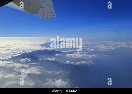 Atemberaubende Luftlandschaftsansicht aus einem Flugzeugfenster, die Berggipfel zeigt, die über einem Meer aus weißen Wolken unter einem klaren blauen Himmelshorizont aufragen Stockfoto