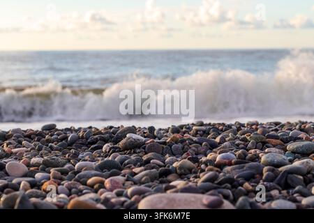 Flachfokusansicht von mehrfarbigen Strandkieseln mit einer im Hintergrund verschwommenen brechenden Meereswelle. Stockfoto